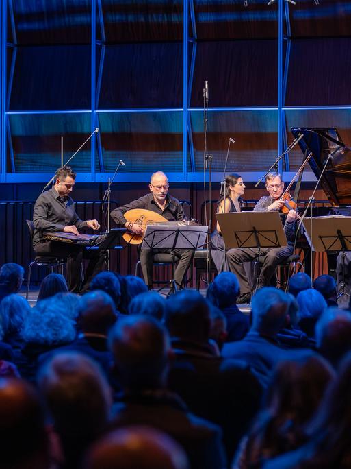 Ein Kanun-Spieler, ein Oud-Musiker, ein Klavierduo, ein Streichquartett und ein Kontrabassist musizieren auf einer blau erleuchteten Bühne vor Publikum
