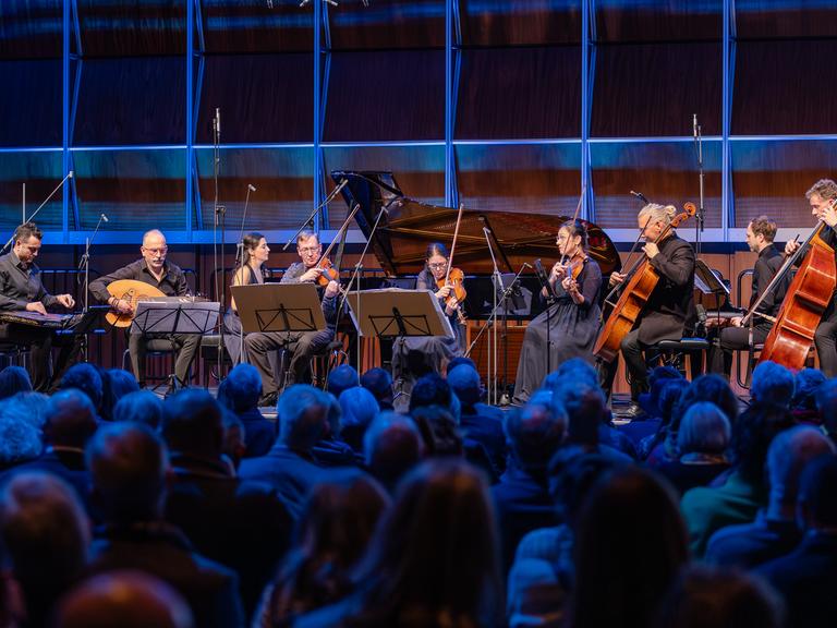 Ein Kanun-Spieler, ein Oud-Musiker, ein Klavierduo, ein Streichquartett und ein Kontrabassist musizieren auf einer blau erleuchteten Bühne vor Publikum