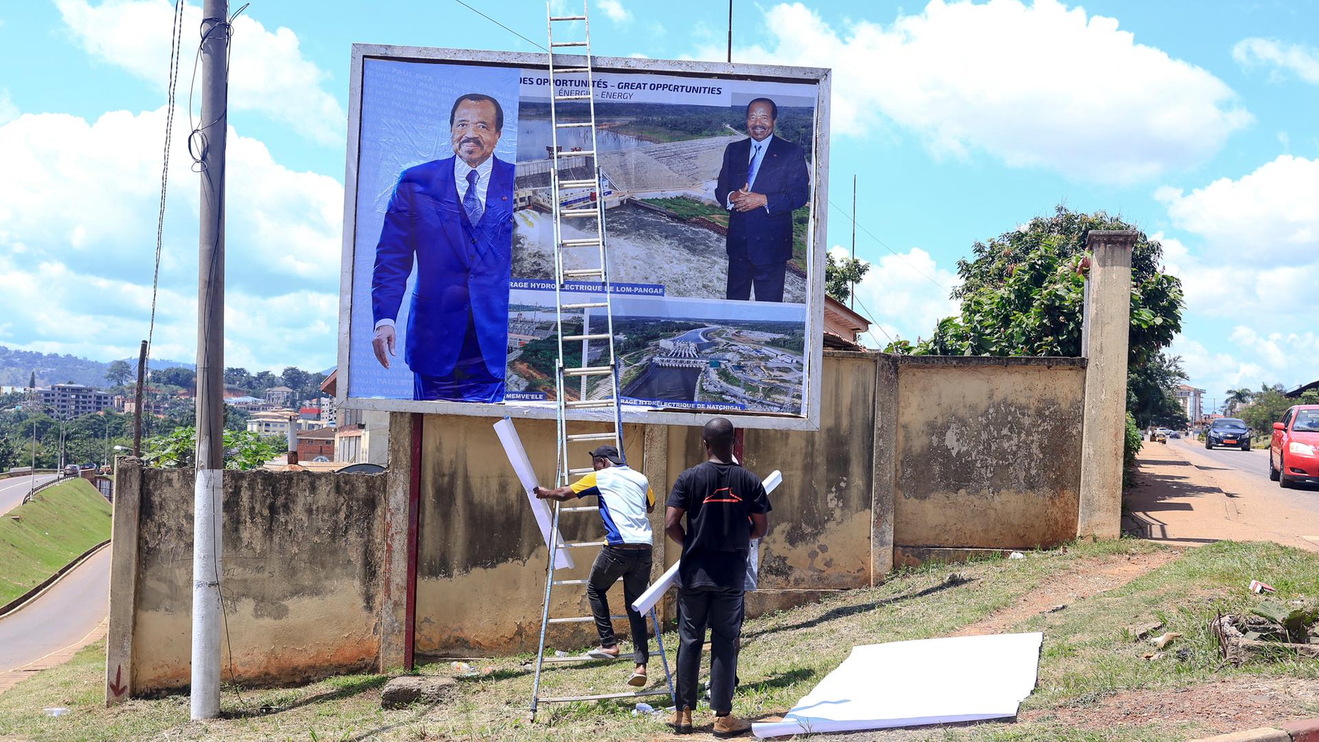 Arbeiter hängen ein Plakat mit dem Gesicht von Präsident Paul Biya an einer Mauer auf.