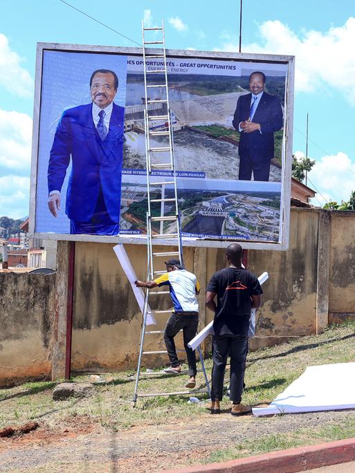 Arbeiter hängen ein Plakat mit dem Gesicht von Präsident Paul Biya an einer Mauer auf.