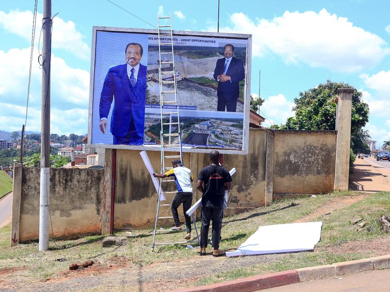 Arbeiter hängen ein Plakat mit dem Gesicht von Präsident Paul Biya an einer Mauer auf.