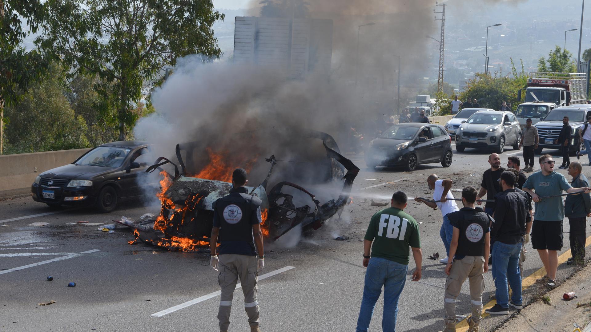 Libanon, Jiyeh: Sicherheitskräfte und Rettungskräfte versammeln sich um ein brennendes Fahrzeug am Schauplatz eines israelischen Luftangriffs, bei dem ein Auto getroffen wurde. 