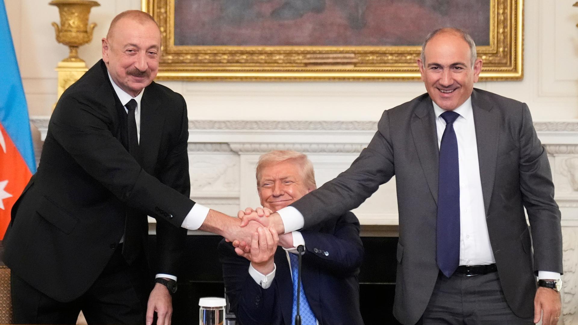 Alijew und Paschinjan stehen nebeneinander, lächeln in die Kamera und geben sich die Hand. Hinter ihnen sitzt der ebenfalls lächelnde Trump und beteiligt sich am Händedruck. Alijew und Paschinjan stehen nebeneinander, lächeln in die Kamera und geben sich die Hand. Hinter ihnen sitzt der ebenfalls lächelnde Trump und beteiligt sich am Händedruck.