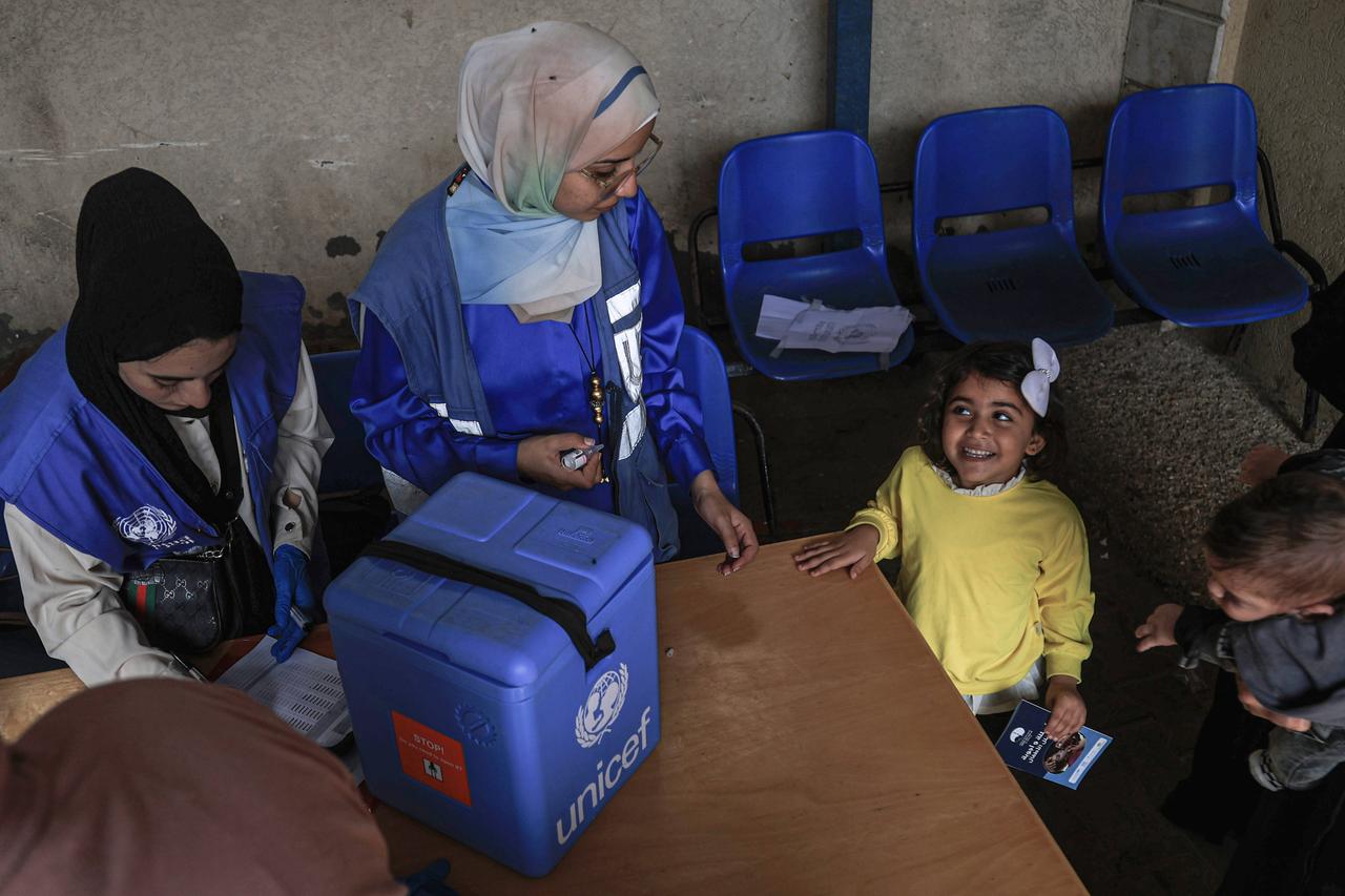 Zwei Frauen mit Kopftüchern und Westen mit dem Emblem der UNWRA darauf stehen an einem Tisch. Vor ihnen steht ein blauer Kühlkoffer mit der Aufschrift Unicef. Neben ihnen steht ein lachendes kleines Kind.