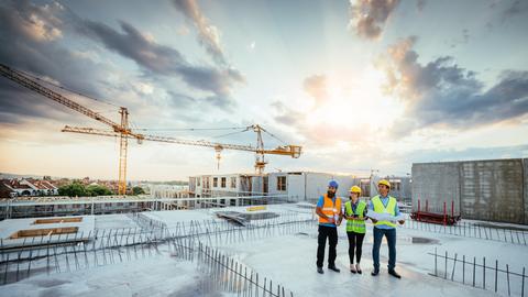Drei Personen in Warnwesten stehen auf dem Dach einer Baustelle. Hinter ihnen geht die Sonne auf.