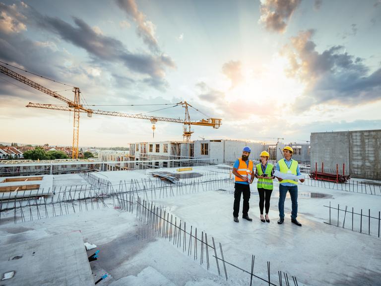 Drei Personen in Warnwesten stehen auf dem Dach einer Baustelle. Hinter ihnen geht die Sonne auf.