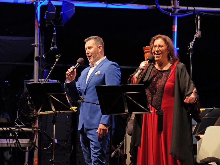 Ein Mann im blauen Anzug und eine Frau in einem schwarz-roten Kleid stehen auf einer Bühne, vor einem Orchester und singen. Sie halten Mikrofone in der Hand.