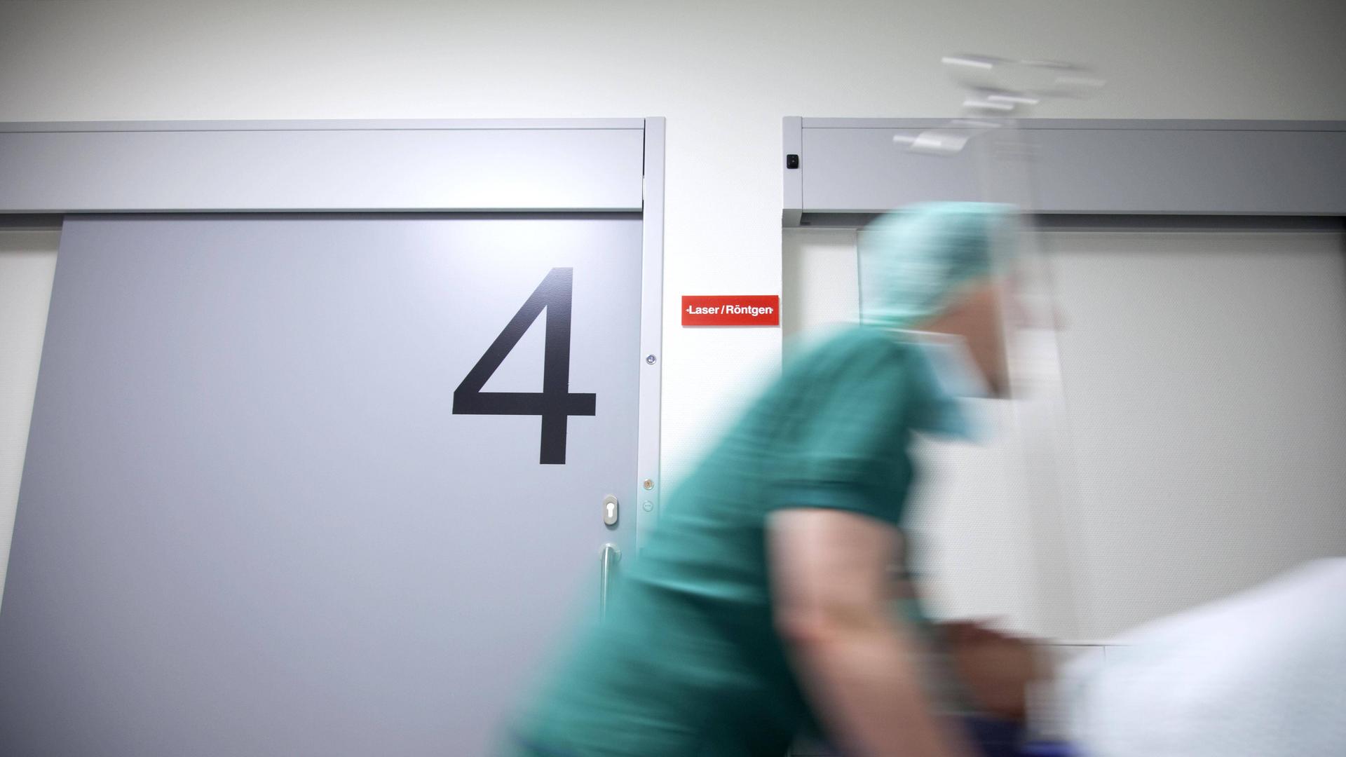 Ein Patient im Bett wird über einen Gang geschoben, im Hintergrund ist eine Tür zum Operationssaal zu sehen.