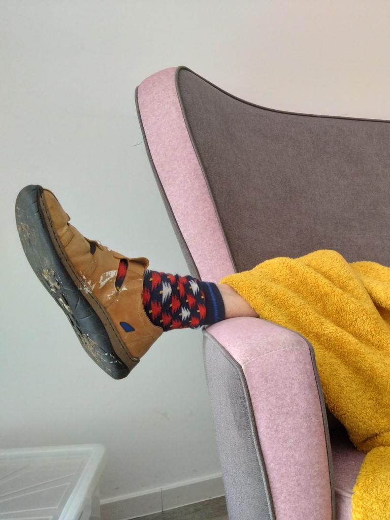Eine Person liegt zugedeckt auf einem Sofa. Unter der Decke schaut ein Fuß heraus, der in einem Hausschuh und bunt gemusterten Socken steckt.