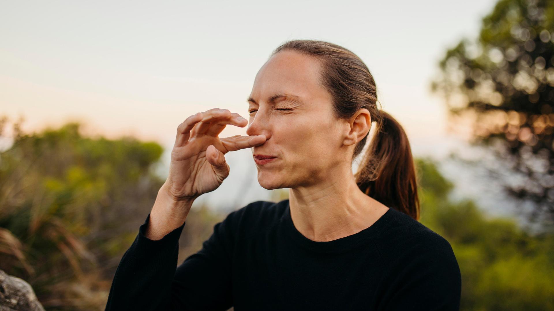 Eine Frau steht mit geschlossenen Augen in der Natur und kratzt sich die Nase, als würde sie ein Niesen unterdrücken.