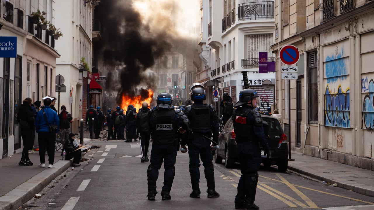 Polizisten stehen in einer Straße, im Hintergrund ist Feuer zu sehen. Links am Straßenrand stehen Passanten.