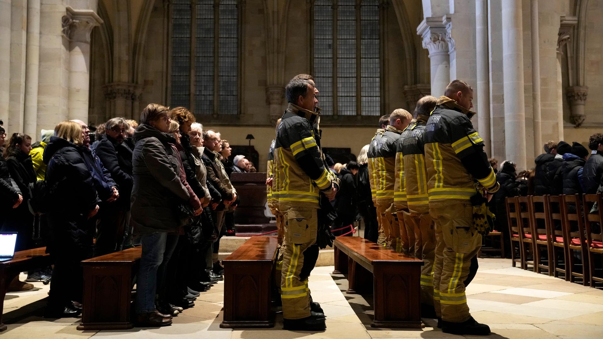 Mehrere Reihen von Kirchenbänken, in denen Trauergäste stehen, darunter Feuerwehrleute.