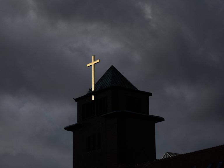 Dunkle Wolken über einem Kirchturm, dessen goldenes Kreuz strahlt.