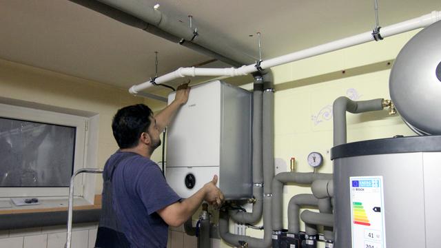 Ein Handwerker der Firma Jänichen Versorgungstechnik befestigt die Abdeckung an einer Steuereinheit der Wärmepumpe im Keller einer Doppelhaushälfte, während rechts die EU-Energieverbrauchskennzeichnung am Pufferspeicher der Wärmepumpe zu sehen ist.