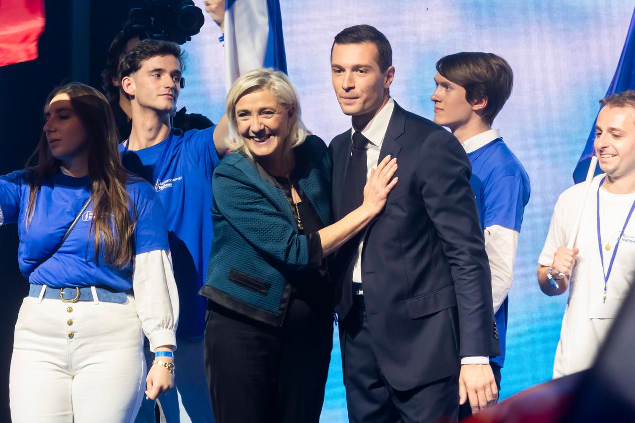 Jordan Bardella und Marine Le Pen stehen auf einer Bühne. Um sie herum stehen vier junge Menschen.