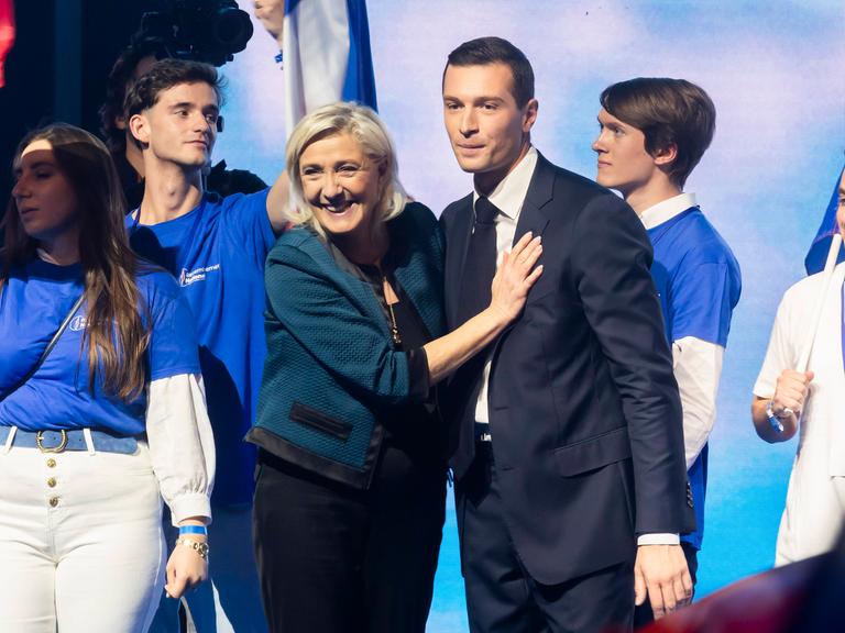 Jordan Bardella und Marine Le Pen stehen auf einer Bühne. Um sie herum stehen vier junge Menschen.
