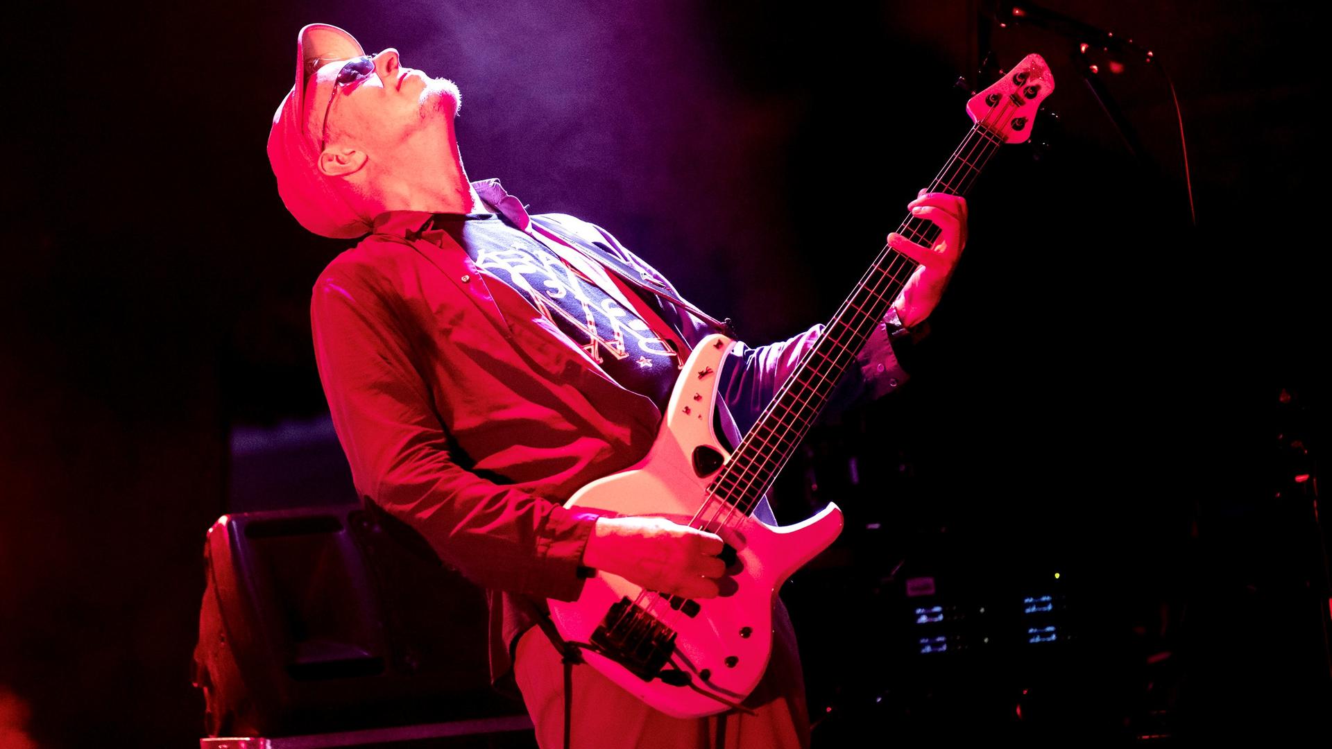 Auf dem Bild ist der Bassist Hellmut Hattler zu sehen, wie er auf einer rot erleuchteten Bühne steht und Bass spielt. 