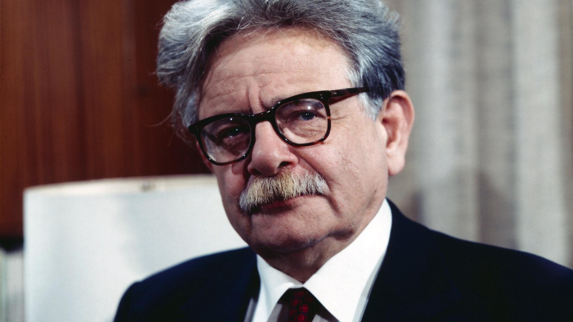 Porträt von Elias Canetti, der eine Brille mit dickem schwarzem Rand und einen schwarzen Anzug mit dunkelroter Krawatte trägt.