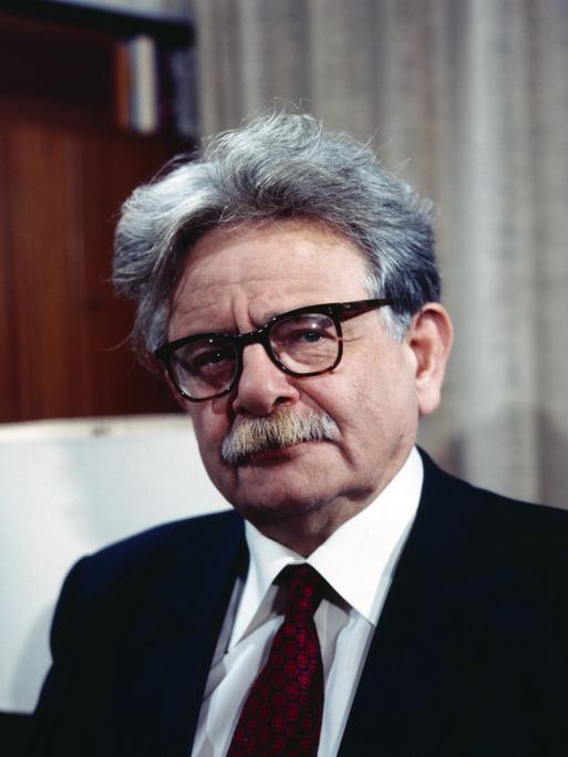 Porträt von Elias Canetti, der eine Brille mit dickem schwarzem Rand und einen schwarzen Anzug mit dunkelroter Krawatte trägt.