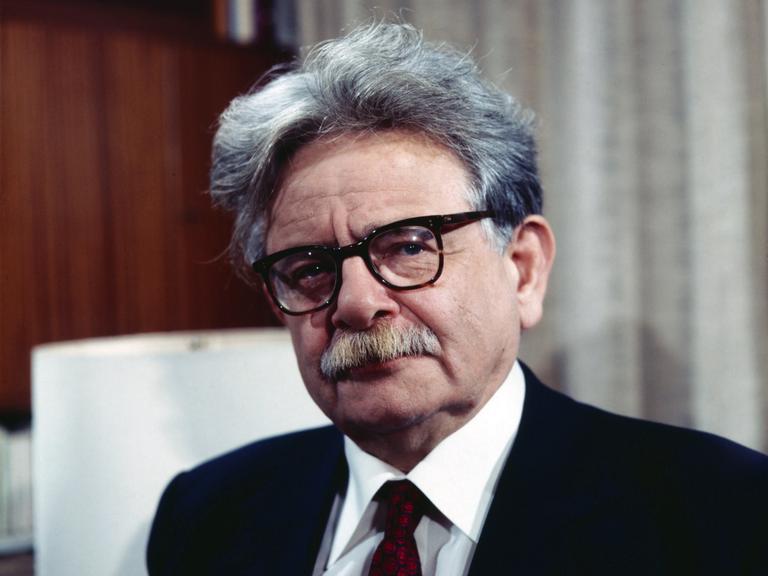 Porträt von Elias Canetti, der eine Brille mit dickem schwarzem Rand und einen schwarzen Anzug mit dunkelroter Krawatte trägt.