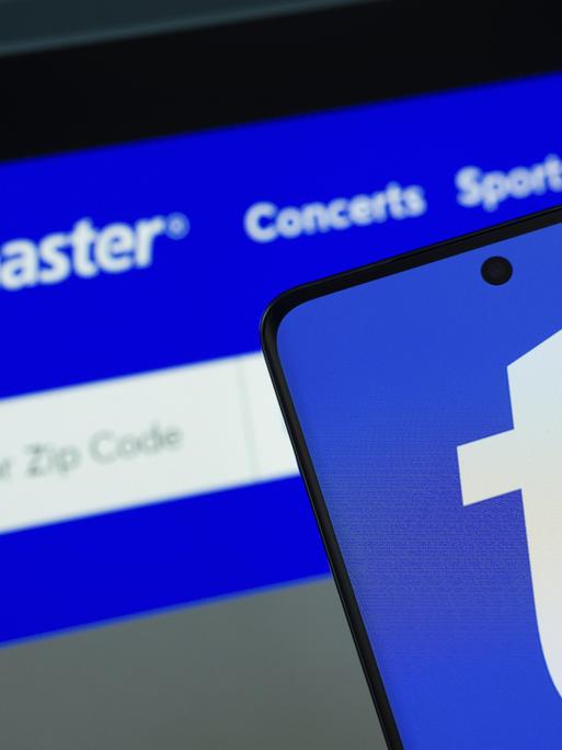 Das Logo von Ticketmaster wird auf einem Smartphone Bildschirm angezeigt, und die Website Ticketmaster ist auf einem Computerbildschirm im Hintergrund zu sehen