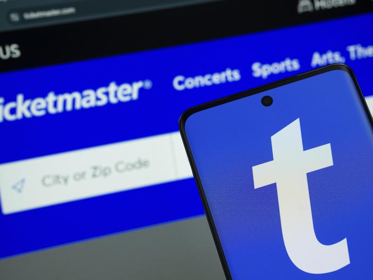 Das Logo von Ticketmaster wird auf einem Smartphone Bildschirm angezeigt, und die Website Ticketmaster ist auf einem Computerbildschirm im Hintergrund zu sehen