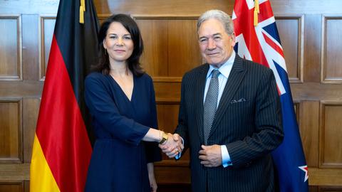 Bundesaußenministerin Annalena Baerbock und der neuseeländische Außenminister Winston Peters geben sich die Hand. Sie stehen vor Flaggen ihrer Länder, dahinter ist eine holzgetäfelte Wand.