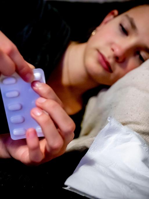 Eine junge Frau liegt im Bett und drückt eine Tablette aus einer Sichtverpackung.