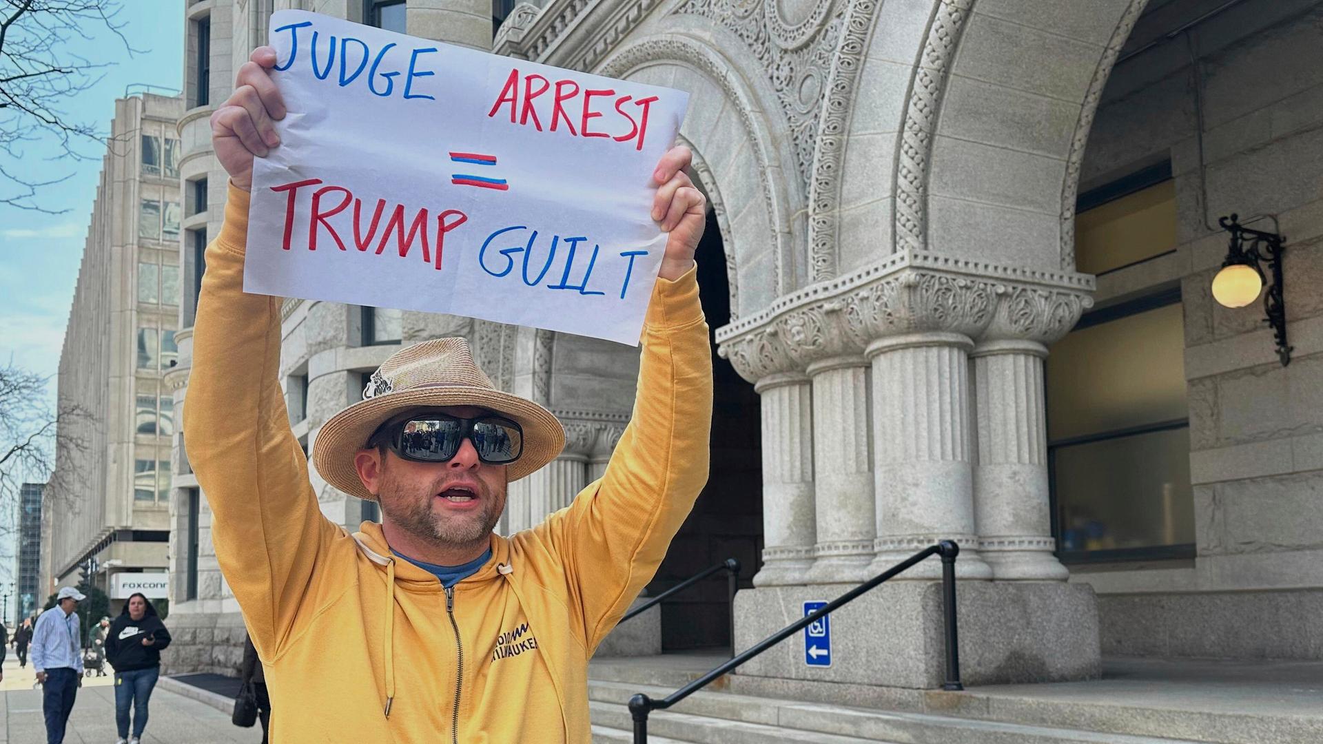 Ein Mann protestiert gegen die zeitweise Festnahme der US-Richterin Hannah Dugan in Milwaukee. Er trägt ein gelbes Sweatshirt und einen Hut, hält ein Plakat hoch, auf dem steht "Judge- Arrest. Trump - Guilt" (Richter festgenommen, Trump schuldig) 

