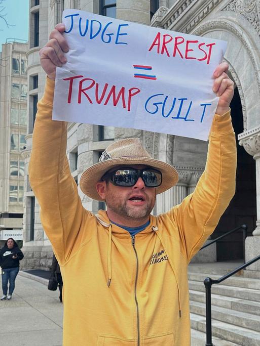 Ein Mann protestiert gegen die zeitweise Festnahme der US-Richterin Hannah Dugan in Milwaukee. Er trägt ein gelbes Sweatshirt und einen Hut, hält ein Plakat hoch, auf dem steht "Judge- Arrest. Trump - Guilt" (Richter festgenommen, Trump schuldig) 

