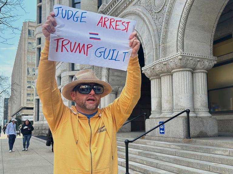 Ein Mann protestiert gegen die zeitweise Festnahme der US-Richterin Hannah Dugan in Milwaukee. Er trägt ein gelbes Sweatshirt und einen Hut, hält ein Plakat hoch, auf dem steht "Judge- Arrest. Trump - Guilt" (Richter festgenommen, Trump schuldig) 

