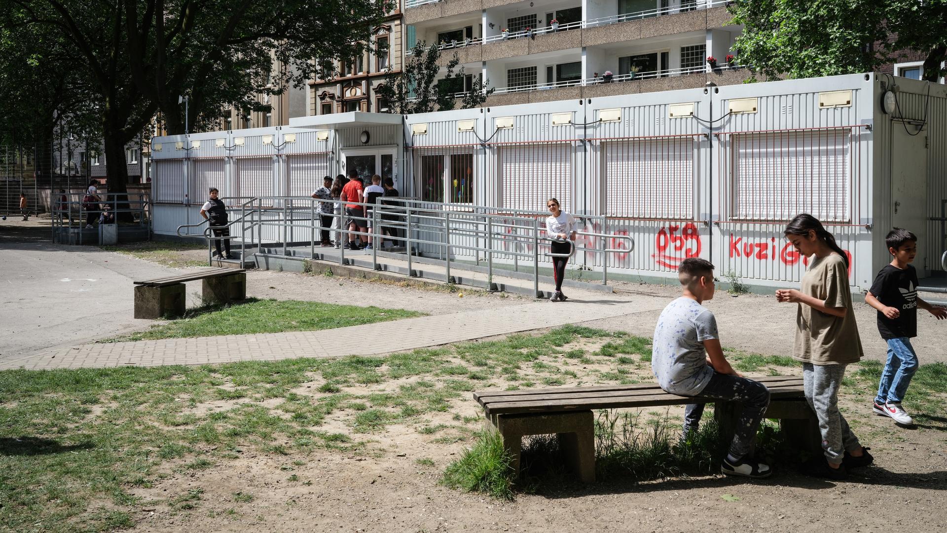 Kinder auf einem Schulhof. Im Hintergrund sind Containerbüros und Plattenbauten zu sehen.