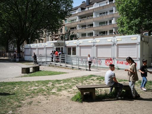 Kinder auf einem Schulhof. Im Hintergrund sind Containerbüros und Plattenbauten zu sehen.