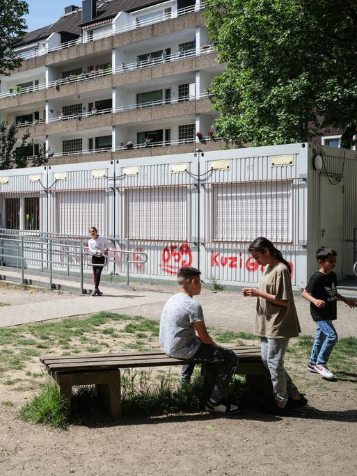 Kinder auf einem Schulhof. Im Hintergrund sind Containerbüros und Plattenbauten zu sehen.