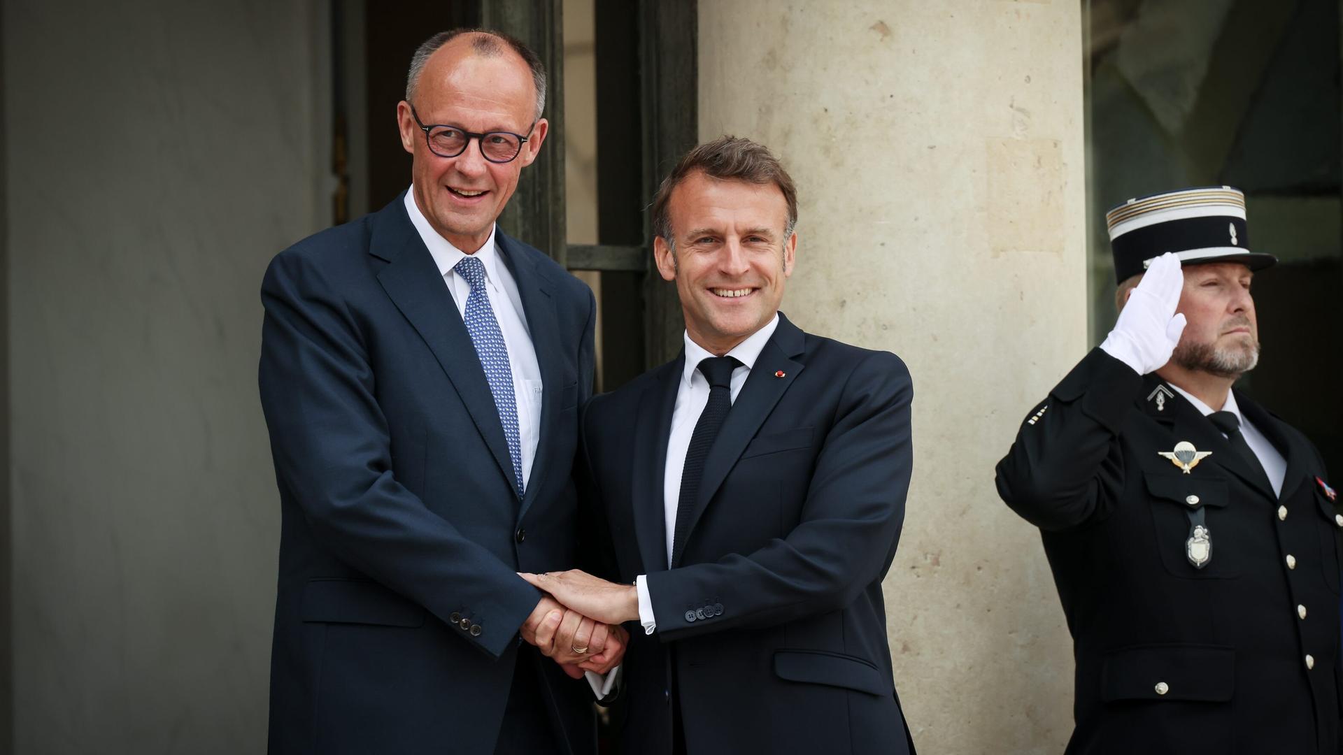 Zwei Männer in Anzügen halten sich an den Händen und lachen in die Kamera: Präsident Emmanuel Macron empfängt Bundeskanzler Friedrich Merz. Zwei Männer in Anzügen halten sich an den Händen und lachen in die Kamera: Präsident Emmanuel Macron empfängt Bundeskanzler Friedrich Merz.