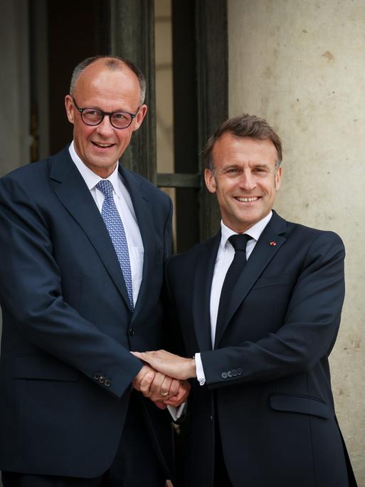 Zwei Männer in Anzügen halten sich an den Händen und lachen in die Kamera: Präsident Emmanuel Macron empfängt Bundeskanzler Friedrich Merz.