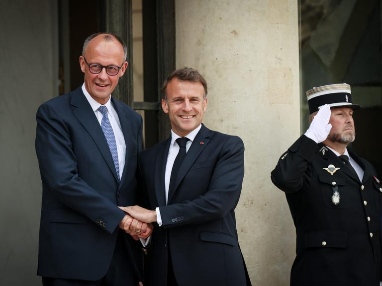 Zwei Männer in Anzügen halten sich an den Händen und lachen in die Kamera: Präsident Emmanuel Macron empfängt Bundeskanzler Friedrich Merz.