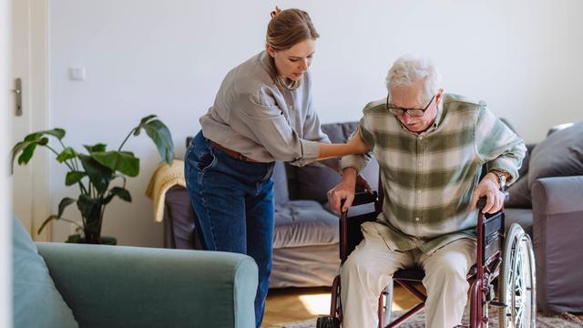 Eine Frau hilft einem alten Mann. Der Mann will sich in einen Rollstuhl setzen. 