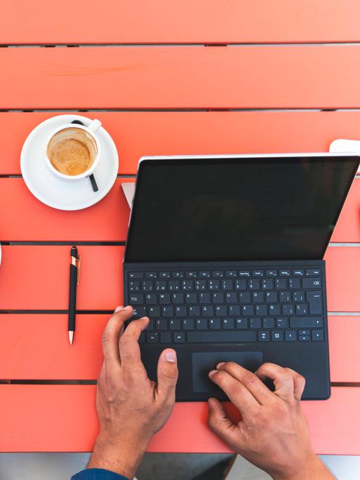 Zwei Personen, von denen man nur die Hände sieht, tippen auf Laptops. Sie stehen auf einem roten Tisch, neben zwei Kaffeetassen.