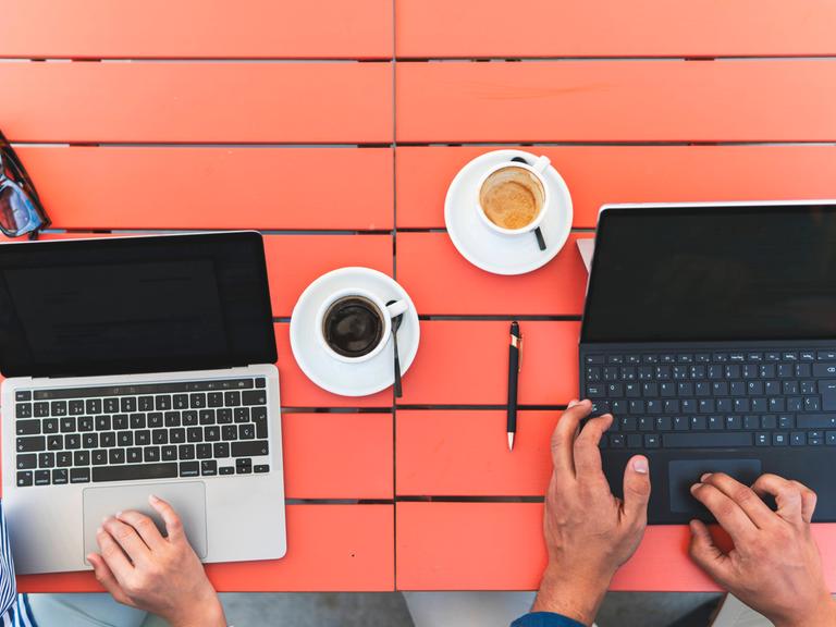 Zwei Personen, von denen man nur die Hände sieht, tippen auf Laptops. Sie stehen auf einem roten Tisch, neben zwei Kaffeetassen.