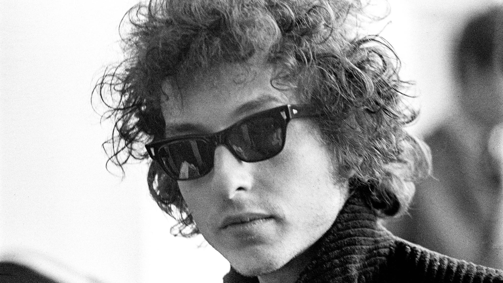 Die Schwarzweißfotografie zeigt den jungen Bob Dylan im Jahr 1966 mit Lockenmähne, Sonnenbrille und Stehkragen.