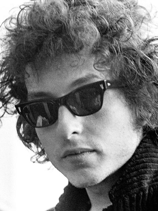 Die Schwarzweißfotografie zeigt den jungen Bob Dylan im Jahr 1966 mit Lockenmähne, Sonnenbrille und Stehkragen.