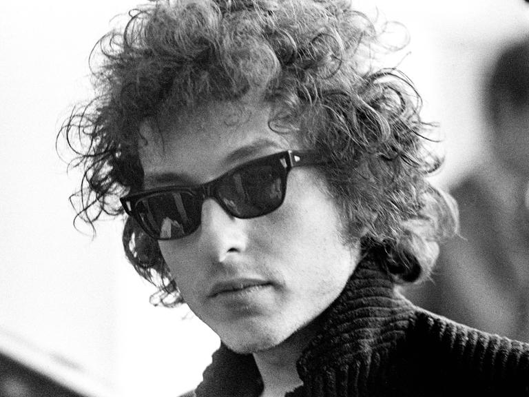 Die Schwarzweißfotografie zeigt den jungen Bob Dylan im Jahr 1966 mit Lockenmähne, Sonnenbrille und Stehkragen.