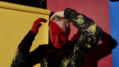 Ein buntes Portrait vor Papierbahnen in rot, gelb und blau. Der Musiker Martin Tingvall streicht sich die blonden Haare zurück, er trägt einen schwarzen Pullover mit wildem, gelbem Muster. Er ist halb im Schatten.
