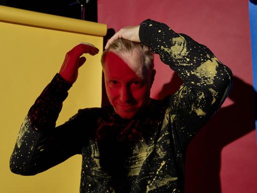 Ein buntes Portrait vor Papierbahnen in rot, gelb und blau. Der Musiker Martin Tingvall streicht sich die blonden Haare zurück, er trägt einen schwarzen Pullover mit wildem, gelbem Muster. Er ist halb im Schatten.