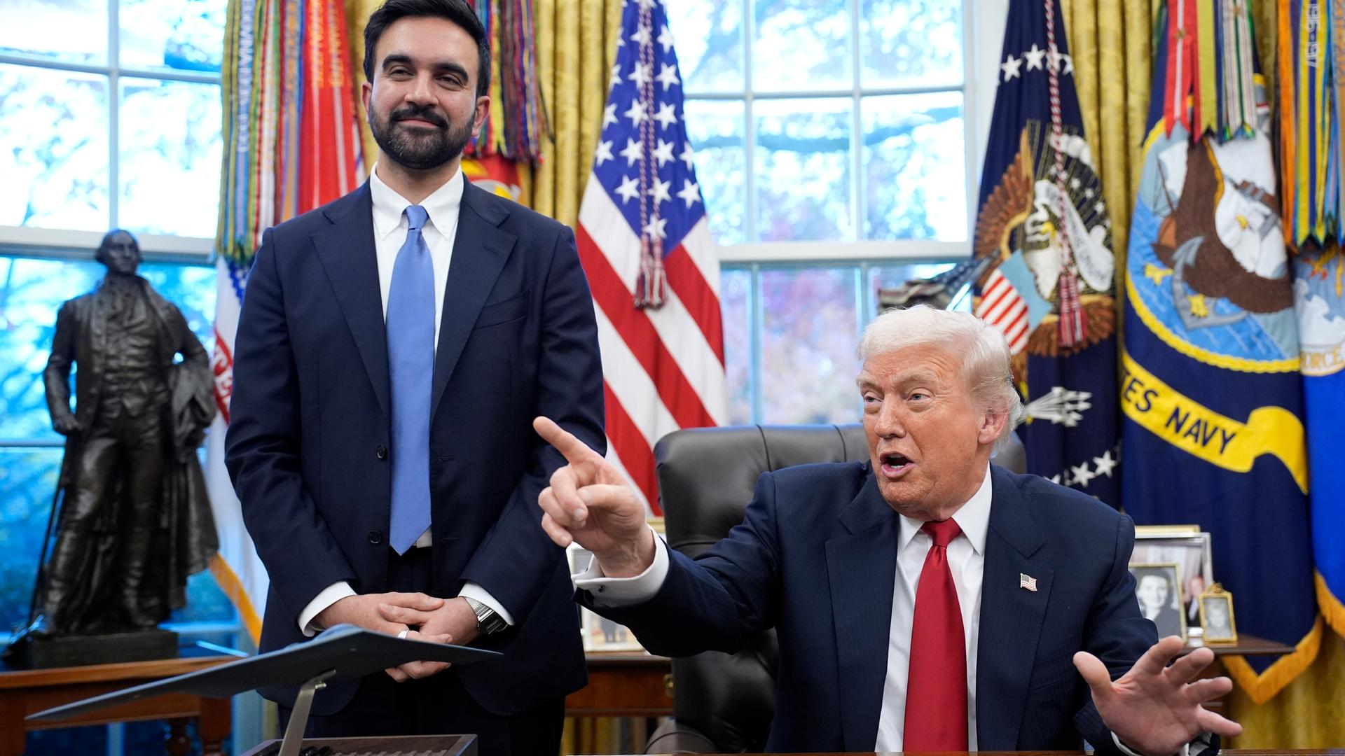 Donald Trump stzt gestikulierend am Schreibtisch im Oval Office. Neben ihm steht Zohran Mamdani und lächelt in die Kamera.