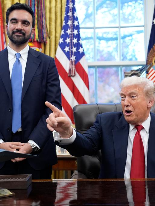 Donald Trump stzt gestikulierend am Schreibtisch im Oval Office. Neben ihm steht Zohran Mamdani und lächelt in die Kamera. Donald Trump stzt gestikulierend am Schreibtisch im Oval Office. Neben ihm steht Zohran Mamdani und lächelt in die Kamera.