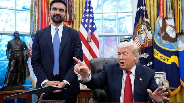 Donald Trump stzt gestikulierend am Schreibtisch im Oval Office. Neben ihm steht Zohran Mamdani und lächelt in die Kamera.