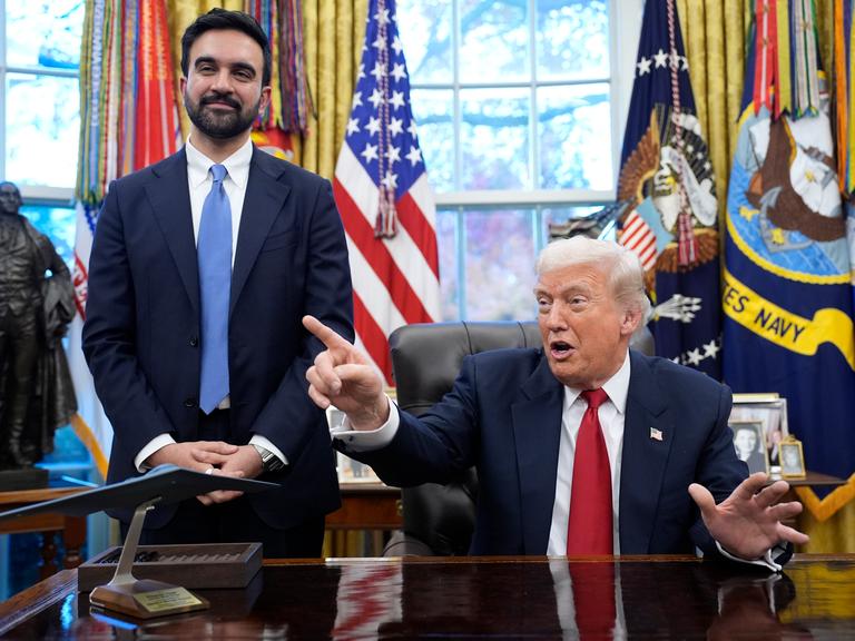 Donald Trump stzt gestikulierend am Schreibtisch im Oval Office. Neben ihm steht Zohran Mamdani und lächelt in die Kamera. Donald Trump stzt gestikulierend am Schreibtisch im Oval Office. Neben ihm steht Zohran Mamdani und lächelt in die Kamera.