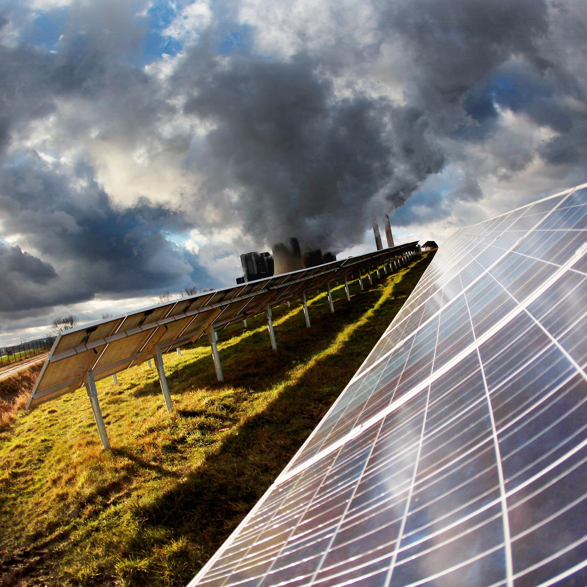 Eine Solaranlage auf der grünen Wiese. Im Hintergrund rauchen die Schlote eines Kraftwerks. 